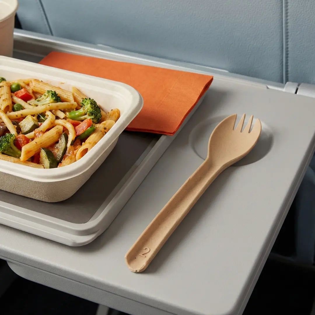CompostEase | Spork on an airplane tray table beside a hot pasta meal, demonstrating durable compostable cutlery for airline service.